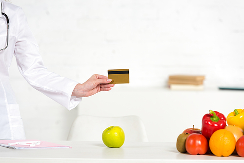 cropped view of dietitian in white coat holding credit card near table with fresh fruits and vegetables