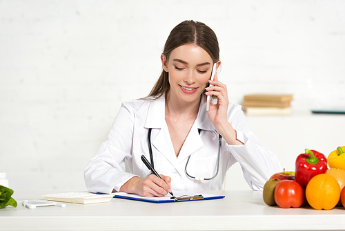 smiling dietitian in white coat talking on smartphone and writing in clipboard at workplace