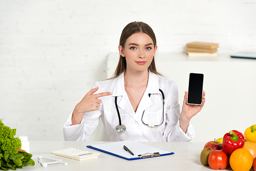 dietitian in white coat holding smartphone with blank screen and pointing with finger at workplace