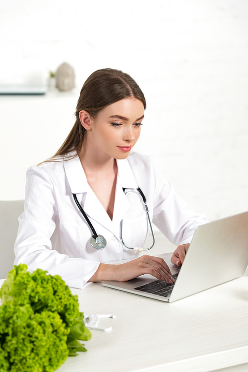 focused dietitian in white coat using laptop at workplace