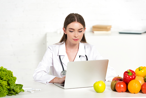focused dietitian in white coat using laptop at workplace with fruits and vegetables