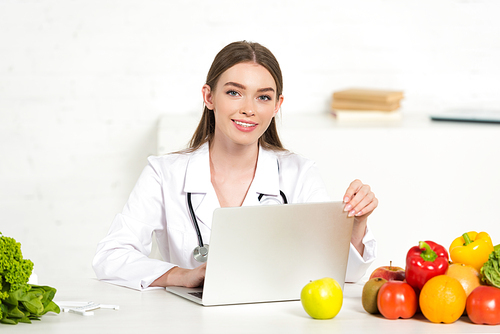 smiling dietitian in white coat using laptop at workplace