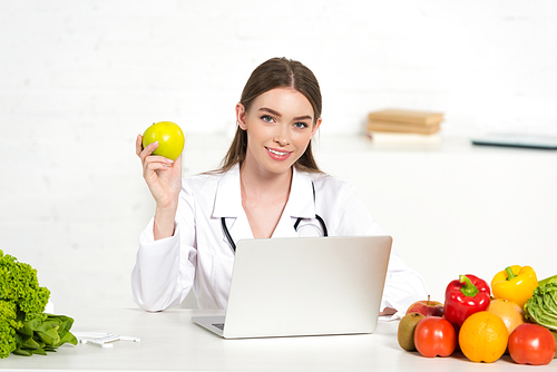 smiling dietitian in white coat holding apple at workplace with laptop