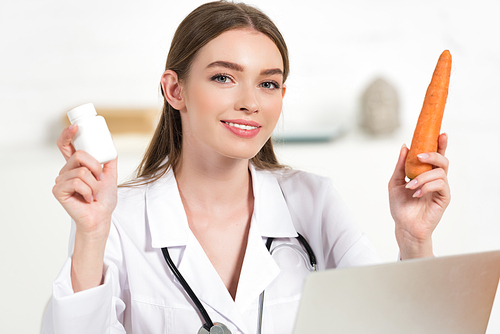 smiling dietitian in white coat holding pills and carrot near laptop