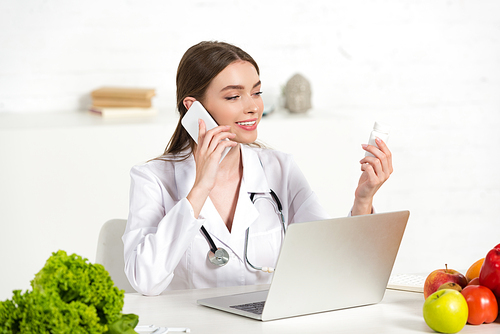 smiling dietitian in white coat talking on smartphone and holding pills at workplace