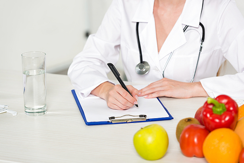 cropped view of dietitian in white coat writing in clipboard at workplace