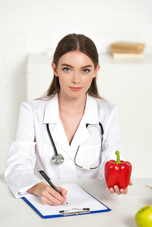 front view of dietitian holding bell pepper and writing in clipboard at workplace