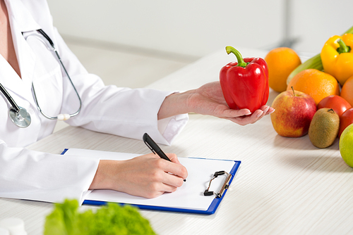partial view of dietitian in white coat holding red bell pepper and writing in clipboard at workplace