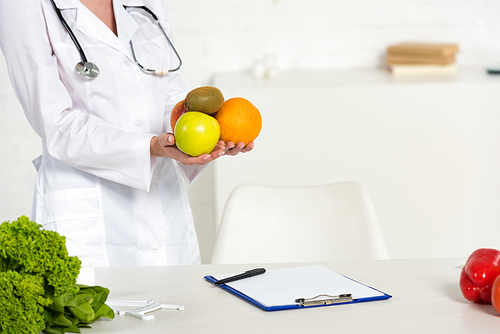 partial view of dietitian in white coat holding fresh fruits near workplace
