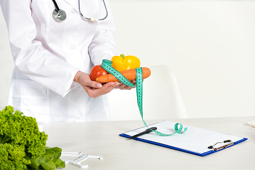 cropped view of dietitian in white coat holding vegetables and measure tape at workplace