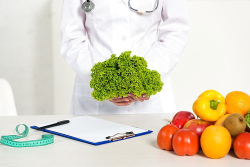 cropped view of dietitian in white coat holding lettuce at workplace