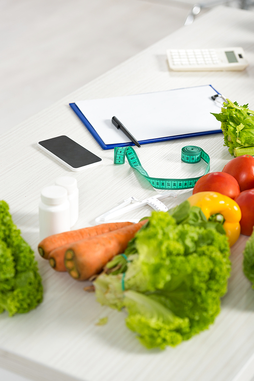 clipboard with pen, measure tape, smartphone with blank screen, calculator, medicine and fresh vegetables on table