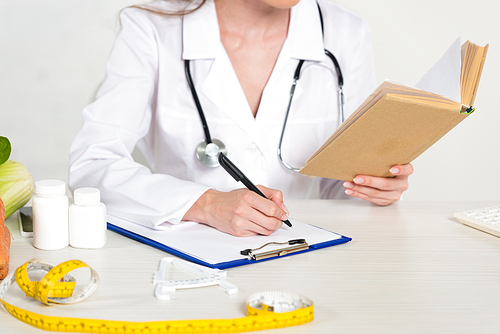 cropped view of dietitian in white coat reading book and writing in clipboard at workplace