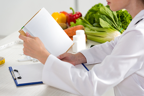 cropped view of dietitian in white coat reading book at workplace
