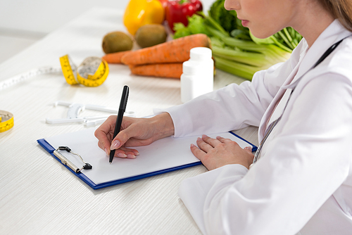 cropped view of dietitian in white coat writing in clipboard at workplace