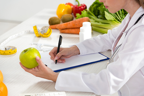 cropped view of dietitian in white coat holding green apple and writing in clipboard