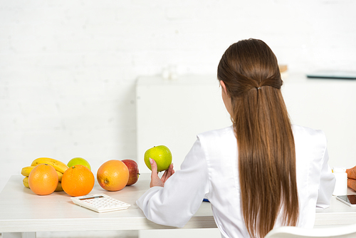 back view of dietitian in white coat holding green apple at workplace