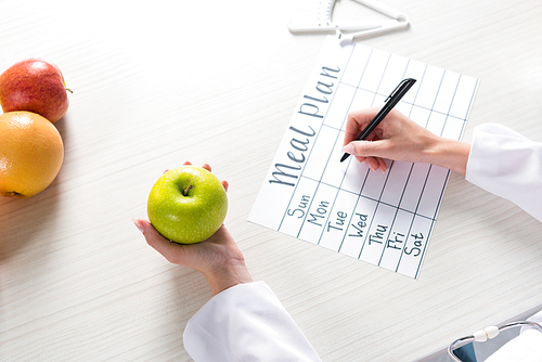 cropped view of dietitian holding apple and writing in meal plan at workplace