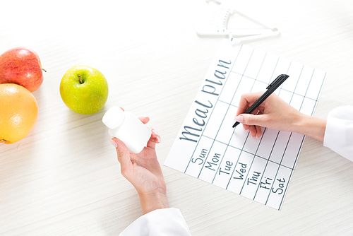 cropped view of dietitian holding pills and writing in meal plan at workplace