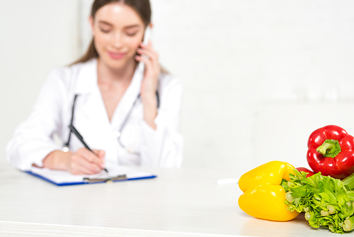 selective focus of dietitian in white coat writing in clipboard and talking on smartphone at workplace