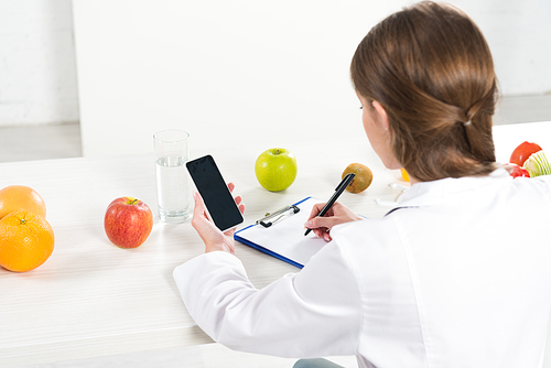 back view of dietitian holding smartphone with blank screen and writing in clipboard at table