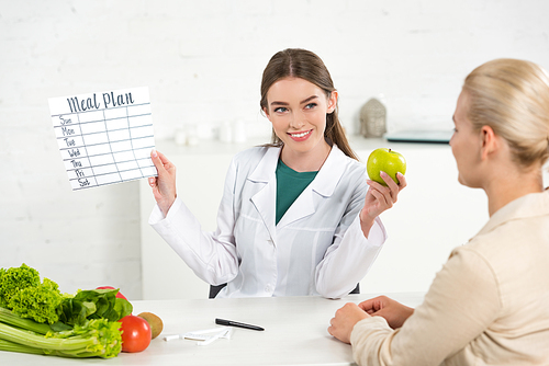 smiling dietitian in white coat holding meal plan and apple and patient at table