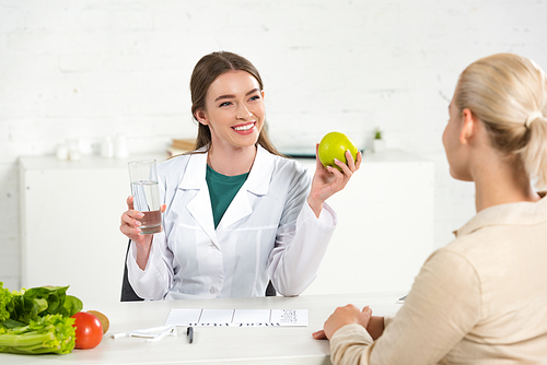 smiling dietitian in white coat holding apple and glass of water and patient at table