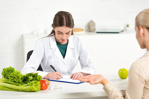 focused dietitian in white coat writing in clipboard and patient at table