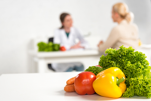 selective focus of dietitian in white coat and patient at table and fresh vegetables on foreground