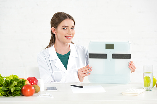 smiling dietitian in white coat holding digital scales at workplace