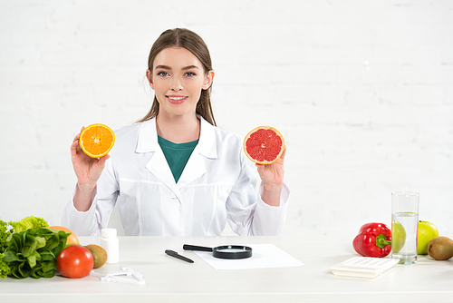 front view of smiling dietitian in white coat holding cut orange and grapefruit