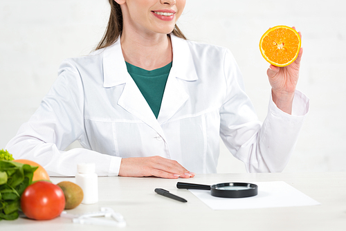 cropped view of smiling dietitian in white coat holding cut orange