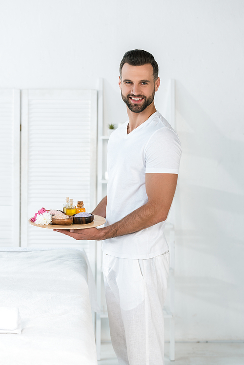 happy bearded man holding tray with bottles of oil, bowls with 씨솔트 and flowers in spa center