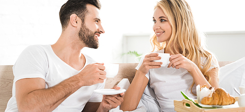 panoramic shot of beautiful couple with cups during breakfast in bed in morning