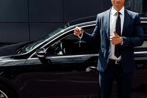 cropped view of african american man in suit holding car key  and showing thumb up near black car