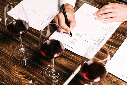 cropped view of sommelier writing in documents at table with wine glasses