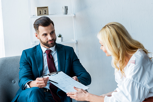 selective focus of handsome recruiter holding clipboard and pen while sitting in armchair near blonde employee