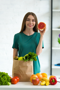 smiling young woman standing near paper bag with fresh vegetables and fruits and holding cut grapefruit at home