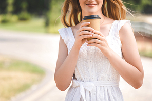 cropped view of young girl in white dress and straw hat smiling and holding paper coffee cup