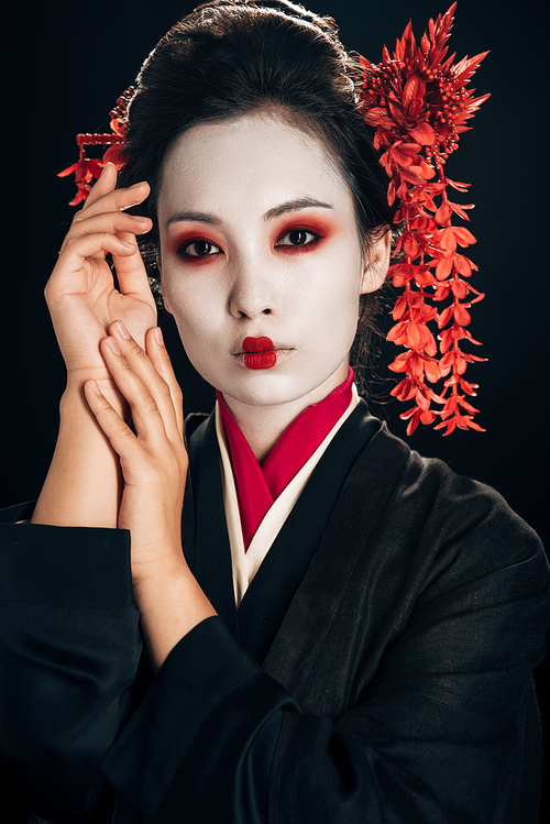 beautiful geisha in black and red kimono and flowers in hair with hands near face isolated on black