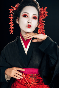 beautiful geisha in black and red kimono and flowers in hair touching chin isolated on black