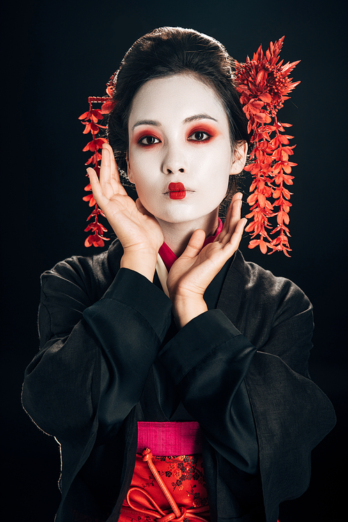 geisha in black and red kimono and flowers in hair with hand near face isolated on black