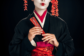 partial view of geisha in black kimono with red flowers in hair holding traditional hand fan isolated on black