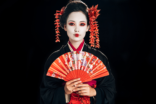 beautiful geisha in black kimono with red flowers in hair holding traditional hand fan isolated on black
