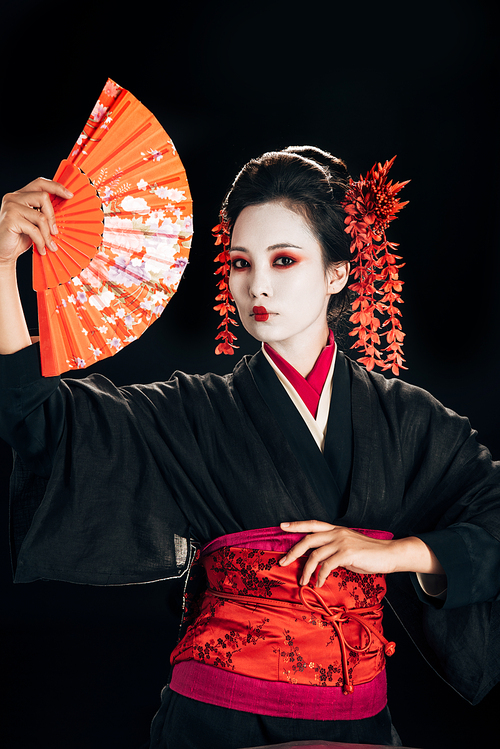 beautiful geisha in black kimono with red flowers in hair posing with traditional hand fan isolated on black