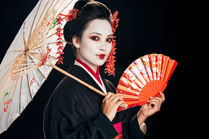 beautiful geisha in black kimono with red flowers in hair holding asian umbrella and hand fan isolated on black