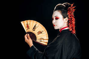 geisha in black kimono with red flowers in hair looking at traditional asian hand fan isolated on black