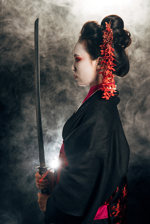 side view of geisha in black kimono holding katana in smoke on black background