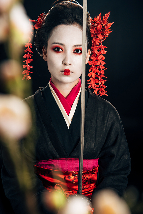 selective focus of confident geisha in black kimono holding katana and sakura branches isolated on black
