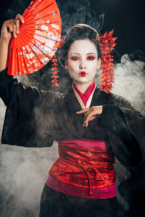 beautiful geisha in black kimono with flowers in hair holding hand fan and gesturing in smoke